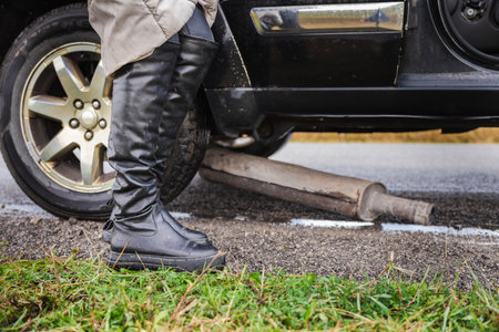 Woman standing near a broken car with a fallen exhaust pipe on the roadsideの写真素材