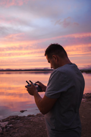 Man operating a drone at sunset near a calm salt lake with vivid orange and pink sky reflections.の写真素材