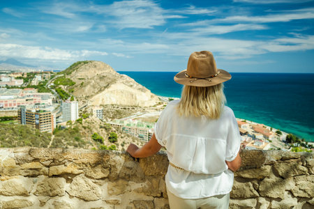 Woman in a hat standing on a stone terrace and looking at the blue sea and the city of Alicante, Spain, on a sunny summer day.の写真素材