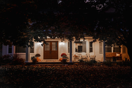 Front porch of an American house illuminated at dusk with autumn leaves on the ground and pumpkins by the door.の写真素材
