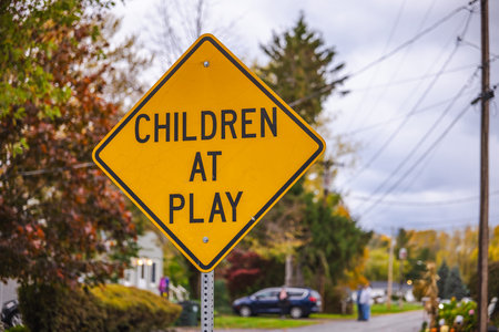 Yellow road sign with the words "Children at play" warning drivers in a residential area surrounded by trees and houses during autumn.の写真素材