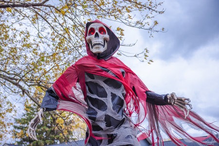 Creepy Halloween skeleton decoration with glowing red eyes and torn red cloak under cloudy autumn sky.の写真素材