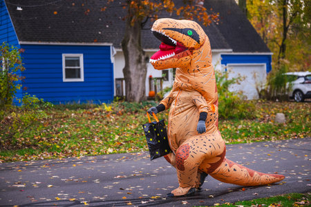 Person in inflatable dinosaur costume walking along suburban street with a Halloween candy bag.
