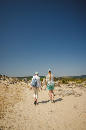 Two women walking hand in hand through the sandy landscape of Pobiti Kamani in Bulgaria under a clear blue sky.の写真素材