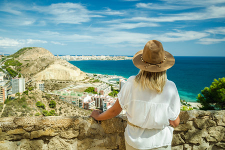 Woman in a hat looking at the blue sea and city from the viewpoint in Alicante, Spain, on a sunny summer day.の写真素材