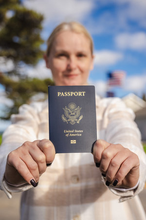 Woman holding a United States passport at arms length toward the camera on a sunny day, with a blurred American flag and suburban houses in the background.の写真素材