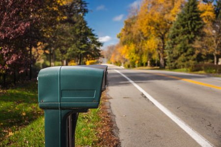 Close-up of a green roadside mailbox on a quiet suburban street during autumn, with colorful trees and soft sunlight in the background.の写真素材