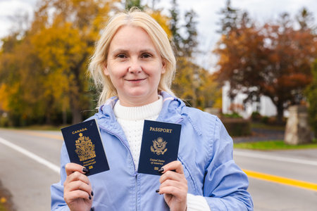 Middle-aged woman standing outdoors in autumn, holding a Canadian passport in one hand and a United States passport in the other near a road.の写真素材