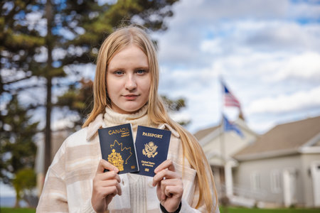 Teenage girl standing outdoors and holding two passports, one Canadian and one US, in front of houses with an American flag in the blurred background.の写真素材