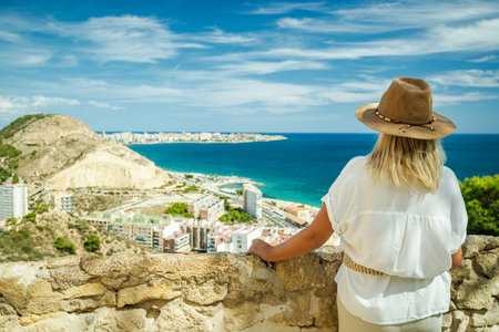 Woman wearing a hat enjoys the sea view and coastline from a scenic viewpoint in Alicante, Spain, on a bright sunny day.の写真素材