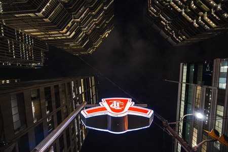 Toronto, Canada, 20 October 2025: A glowing TTC subway entrance sign rises among the towering downtown skyscrapers. The illuminated red emblem of the Torontoのeditorial素材