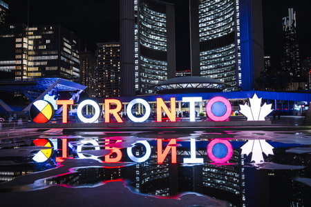 Toronto, Canada, 20 October 2025: Night view of the illuminated Toronto sign at Nathan Phillips Square with colorful reflections on the wet surface and city skyscrapers glowing in the background.のeditorial素材