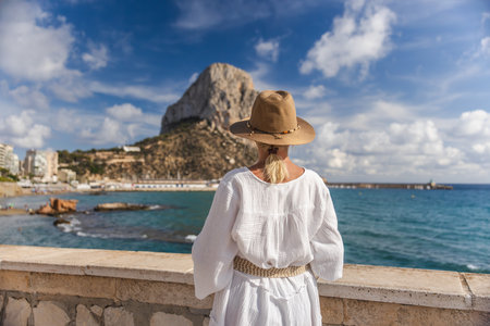 Young woman in a white dress and hat standing by the sea in Calpe, Spain, looking toward the Penon de Ifach rock on a bright sunny dayの写真素材