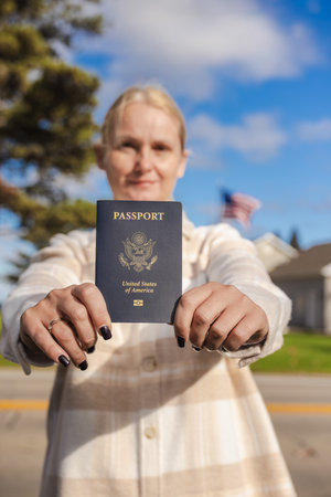 Woman holding a United States passport in both hands toward the camera while standing outdoors, with houses, trees and a blue sky in the background.の写真素材