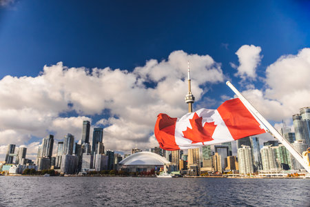Toronto, Canada, 20 October 2025: Canadian flag fluttering above the water with a panoramic view of Torontos downtown skyline in the distance, featuring iconic high-rise buildings and the CN Tower under bright clouds.のeditorial素材