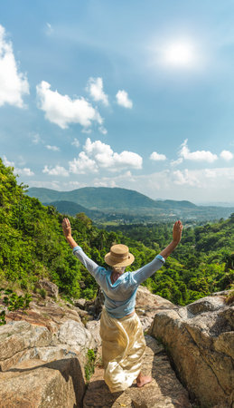 Young woman standing on rocky viewpoint with raised arms overlooking lush tropical valley and...