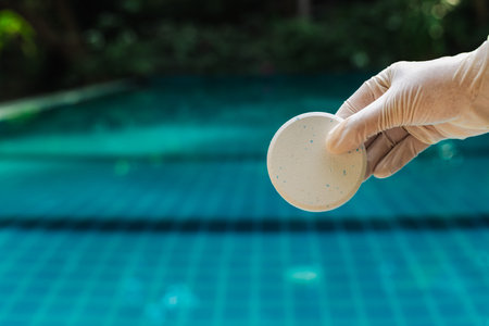 Gloved hand holding chlorine tablet over swimming pool water with blue tiles and soft blurred...