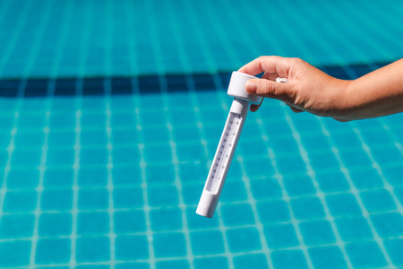 Hand holding pool thermometer measuring water temperature during swimming pool maintenance with...