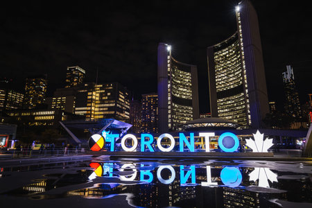 Night view of illuminated Toronto sign with city skyline and reflections at Nathan Phillips Square.