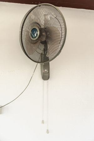 Wall mounted electric fan with pull cords on white wall, household cooling device providing air...