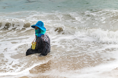 Ko Samui, Thailand, 16 March 2026: Local woman collecting small edible clams in shallow sea waves on...