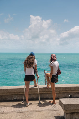 Two young women standing on seaside promenade looking at turquoise oceanの写真素材