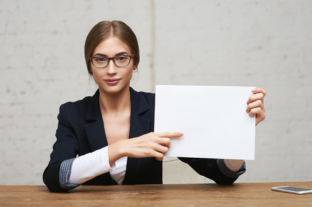 Portrait of young business woman in glasses sitting in office and showing blank white paperの写真素材