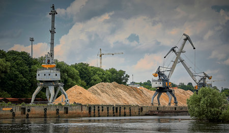 River port in winter. Port cranes on the bank of the river. Embankment of the river port, a barge at...