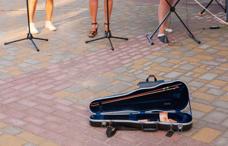 The image shows an open violin case on the ground near the musicians. There is money (tips) in the case. The case is black and made of hard plastic. The musicians are standing in the background.の写真素材