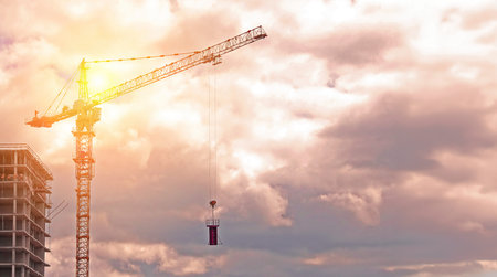 A construction crane stands tall against a backdrop of soft pink and white clouds, with a building...
