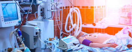 A patient lies in a hospital bed, connected to life support equipment. The room is brightly lit,...