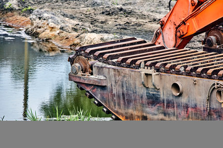 A close-up of a rusted dredge, its treads and hull resting on the bank of a tranquil waterway. The stillness of the water reflects the surrounding landscape.の写真素材