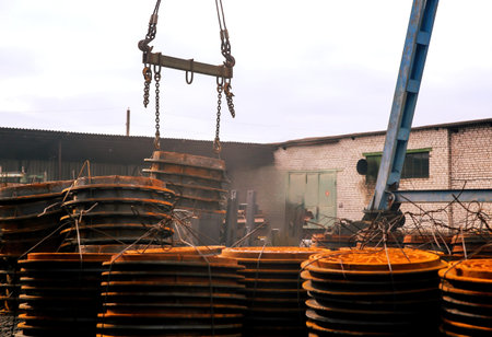 A crane hoists large, circular metal plates at an industrial site. Surrounding stacks of rusty plates indicate ongoing construction during overcast weather.の写真素材
