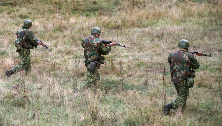 Soldiers in camouflage uniforms patrol a grassy field while carrying rifles during a military...