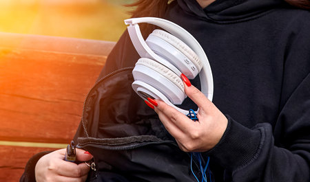 A woman wearing a black hoodie sitting on a bench in a sunny park, holding white over-the-ear headphones with a cord. The background is blurry, creating a serene outdoor setting.の写真素材