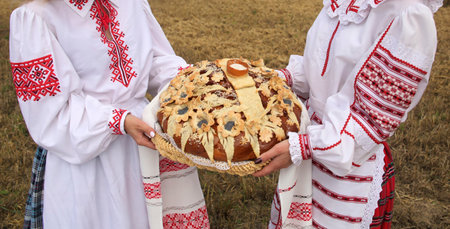 Women in traditional folk costumes present a beautifully decorated bread in a rural field, honoring cultural traditions and festivities.の写真素材