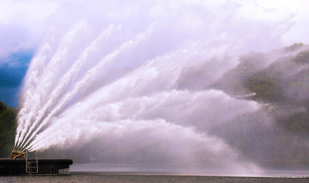 A series of large water jets creates a stunning display at a lakeside, with sunlight shimmering on the water's surface.の写真素材