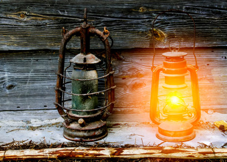 Two vintage lanterns stand beside each other against a wooden backdrop, one glowing warmly while the other retains an aged, weathered appearance.の写真素材