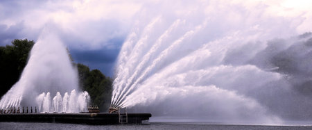 Water jets shoot high into the air from a fountain in a picturesque lake surrounded by lush greenery under a cloudy sky.の写真素材