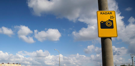 A camera symbol and radar sign are displayed on a pole, set against a backdrop of fluffy clouds and a bright blue sky.の写真素材