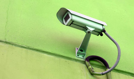 A security camera is mounted high on a bright green wall, monitoring the area in bright daylight,...