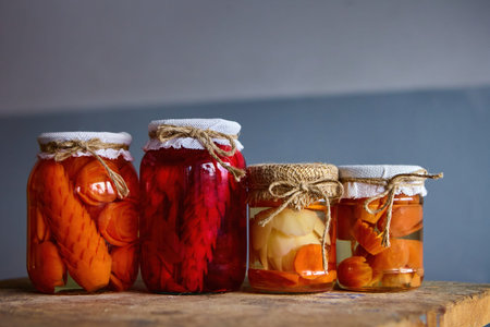 Four jars filled with pickled vegetables display vibrant colors in a rustic kitchen on a wooden table, illuminated by soft light.の写真素材
