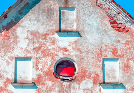 A weathered building wall shows peeling paint and bricks, showcasing a circular window framed by...