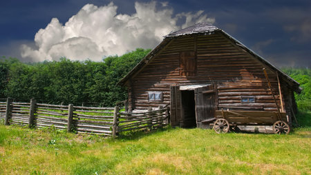 A rustic wooden barn stands in a lush green field, surrounded by trees. Dark clouds loom above, hinting at an impending storm. The tranquil setting reflects pastoral life.の写真素材