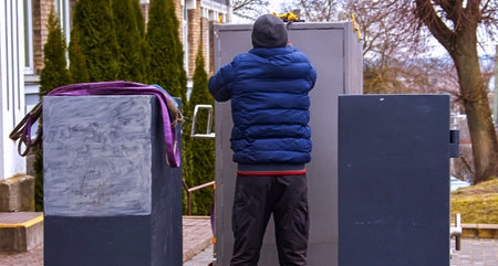 In an outdoor area, an individual in a blue jacket is focusing on adjusting a gray metal structure. The setting features trees and a pathway, indicating a public space.の写真素材