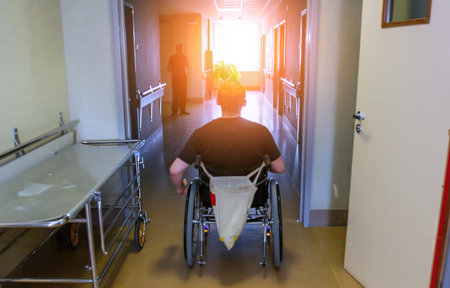 A person in a wheelchair is moving through a hospital corridor, bathed in warm afternoon light, with shadows in the background.の写真素材