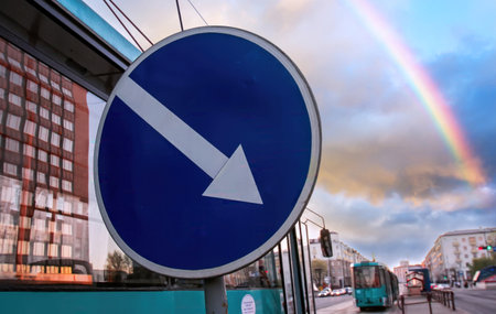 A lively city street features a traffic sign with a downward arrow, set against a backdrop of a rainbow and passing tram under a cloudy sky.の写真素材