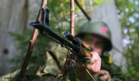 A soldier in full uniform takes aim with a rifle while lying on the ground in a wooded area during a...
