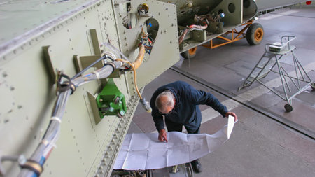 An engineer closely studies technical drawings while inspecting an aircraft in a hangar, ensuring...