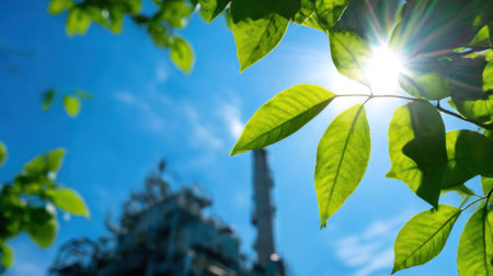 Sunlight filters through vibrant green leaves, highlighting nature, with an industrial structure visible in the background under a blue sky.の素材